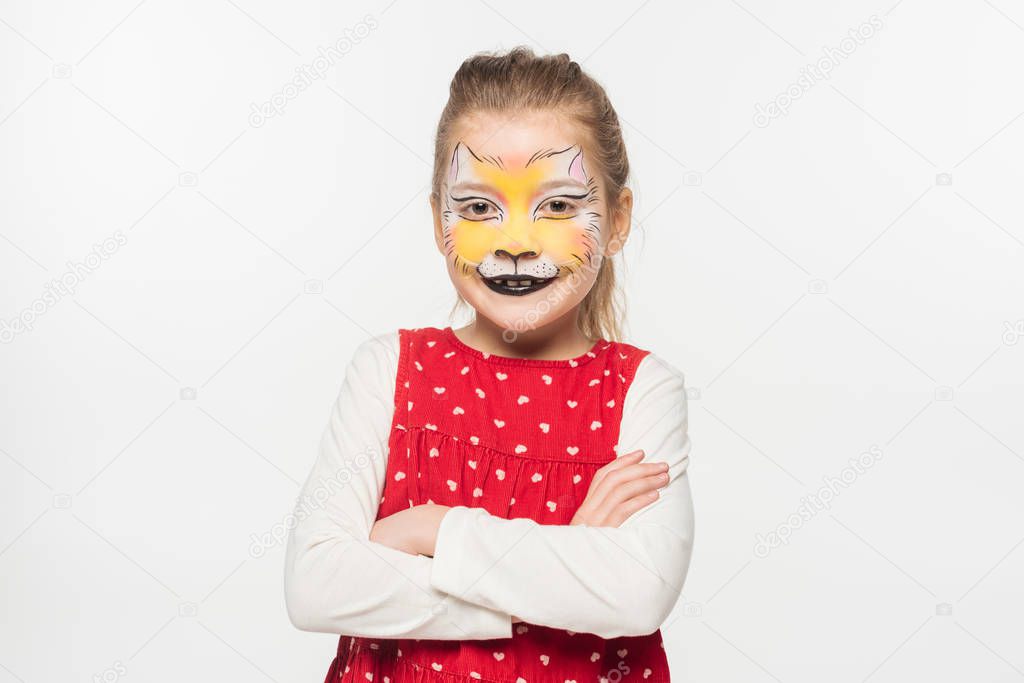 cute kid with tiger muzzle painting on face standing with crossed arms while looking at camera isolated on white