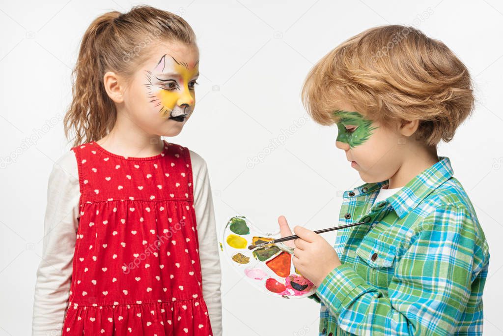 adorable boy with painted gecko mask holding palette and paintbrush near friend with tiger muzzle painting on face isolated on white