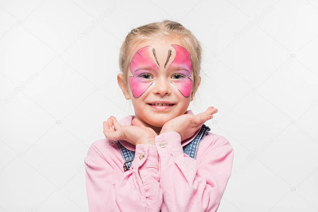 smiling child with butterfly painting on face holding hands near face while looking at camera isolated on white