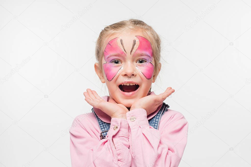 excited child with butterfly painting on face holding hands near face while looking at camera isolated on white