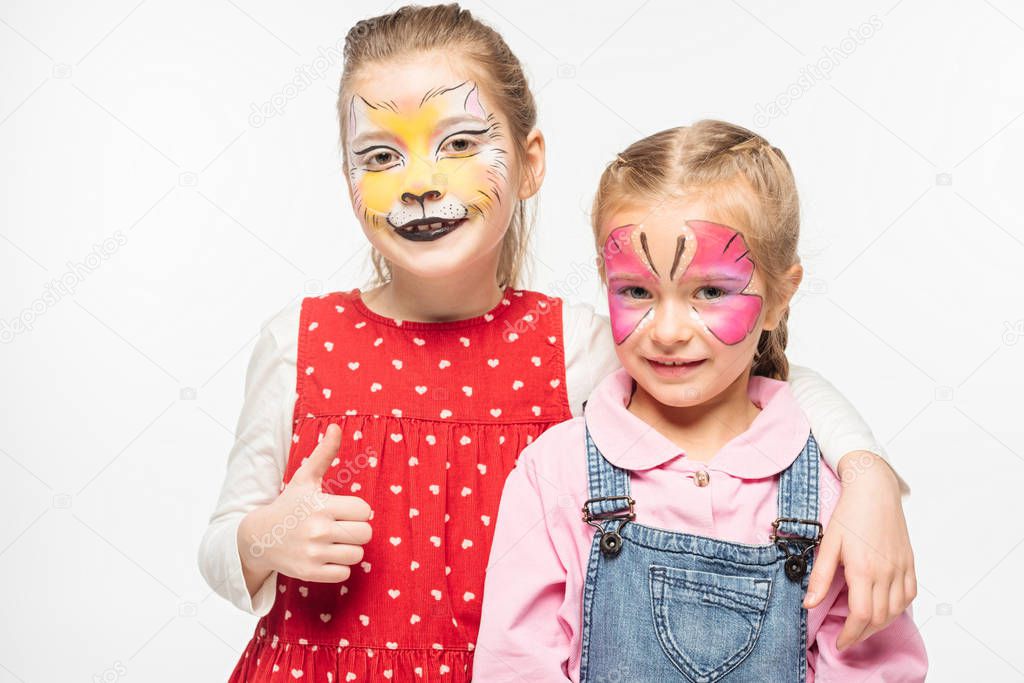adorable kid with cat muzzle painting on face showing thumb up while embracing friend with painted butterfly mask isolated on white