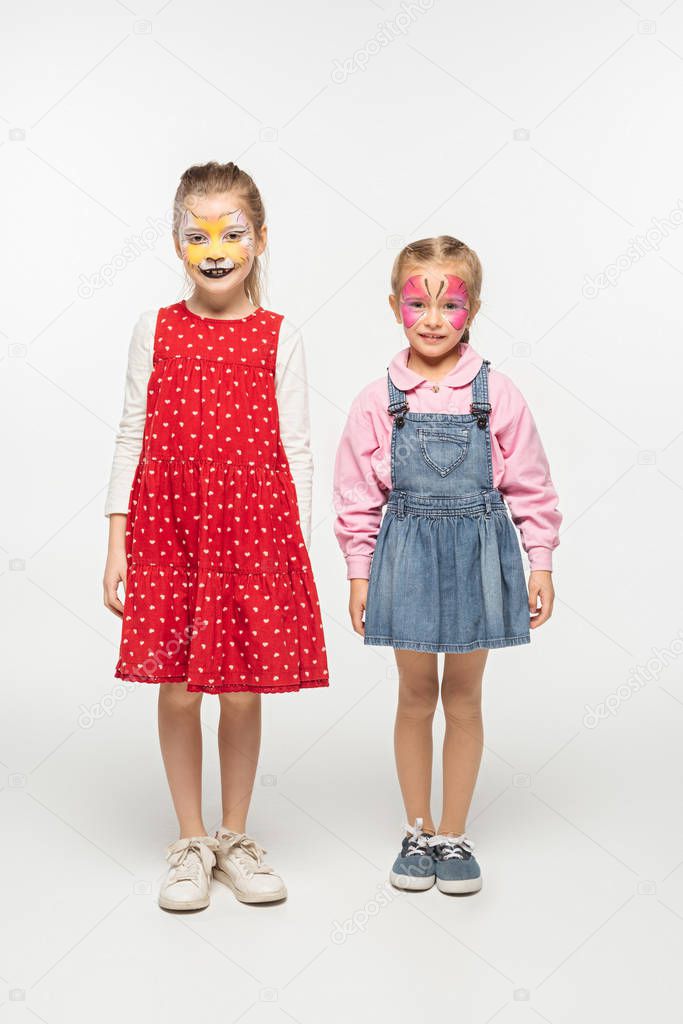 full length view of adorable children with cat muzzle and butterfly paintings on faces standing on white background