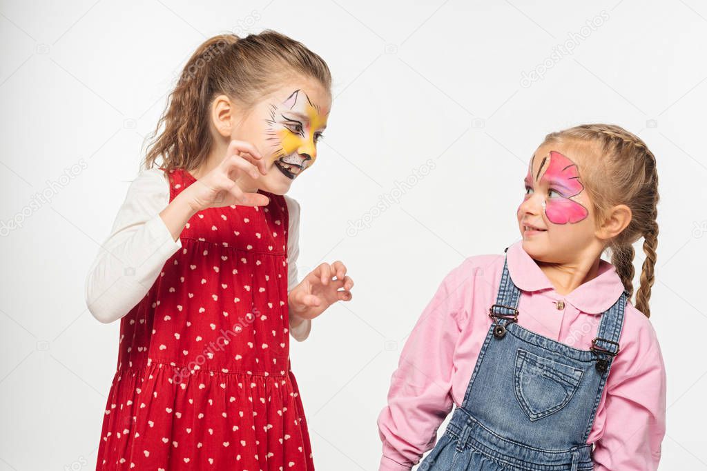cheerful kid with cat muzzle painting on face scaring friend isolated on white