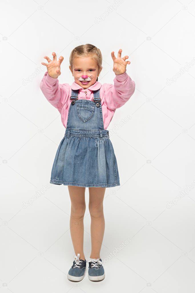 full length view of cute kid with cat muzzle painting on face showing frightening gesture while looking at camera on white background