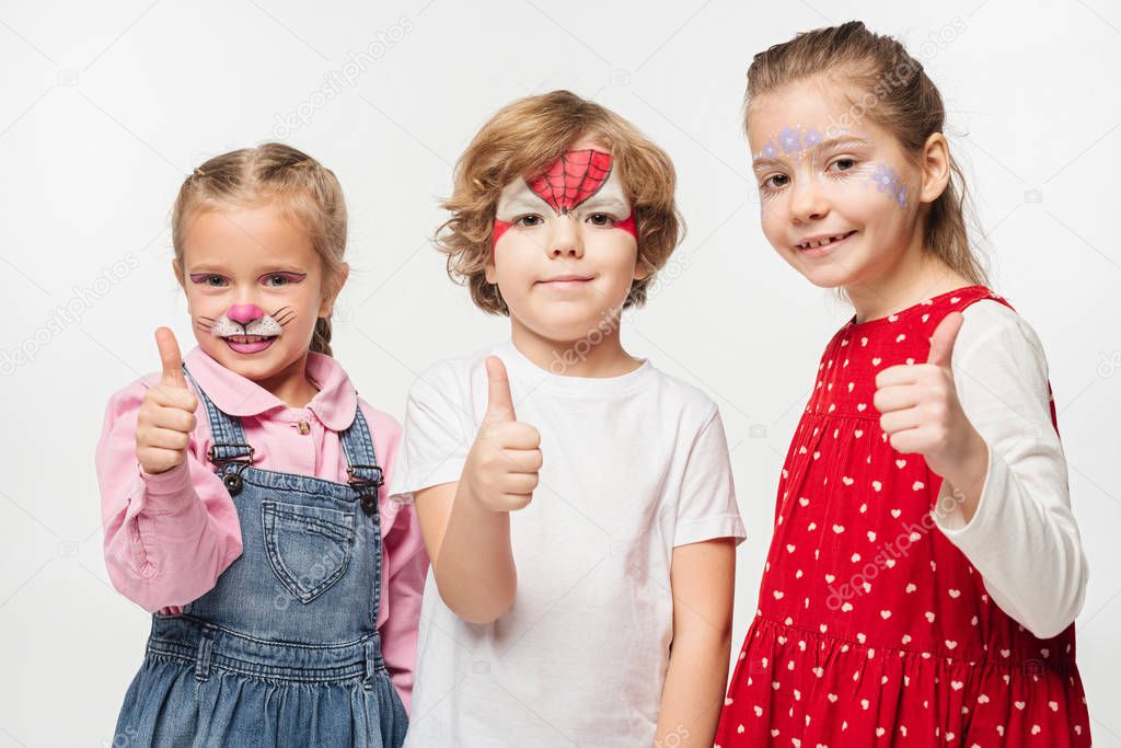 cheerful friends with colorful face paintings showing thumbs up while looking at camera isolated on white