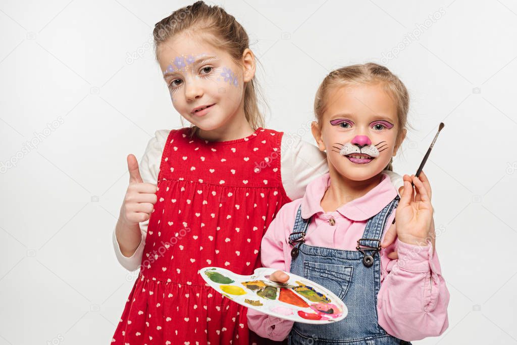 adorable kid with cat muzzle painting of face holding palette and paintbrush, while friend with floral mask showing thumb up isolated on white
