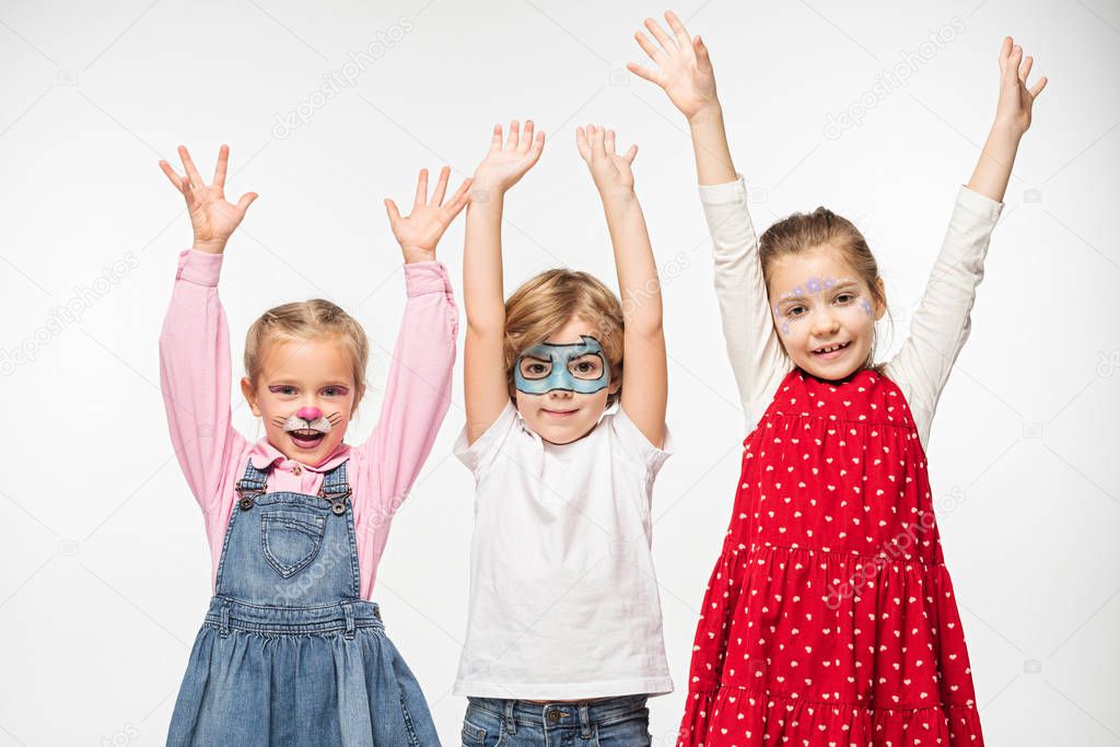 smiling friends with colorful face paintings standing with raised hands while looking at camera isolated on white