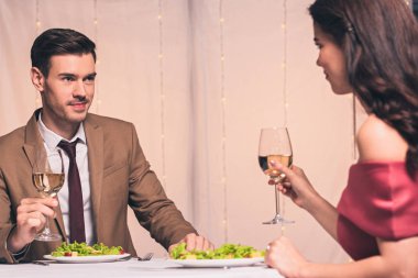 happy, elegant man and woman sitting at served table and holding glassed of white wine clipart