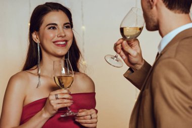 selective focus of attractive, happy girl looking at boyfriend while holding glass of white wine clipart