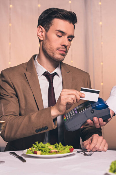 partial view of waiter with payment terminal near handsome man holding credit card while sitting in restaurant