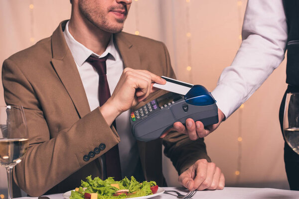 partial view of waiter with payment terminal near man holding credit card while sitting at served table