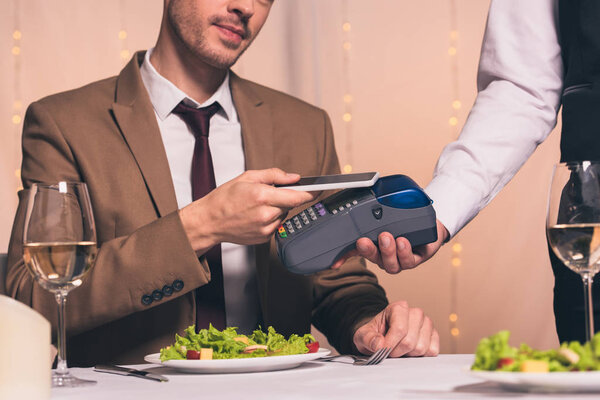 cropped view of waiter with payment terminal near man holding credit card while sitting at served table