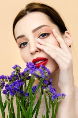 beautiful woman looking at camera and touching face near purple flowers isolated on beige 