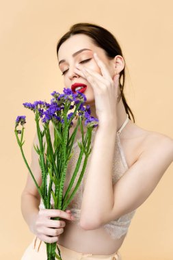 beautiful woman with closed eyes touching face and holding purple limonium flowers isolated on beige 
