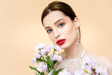 pretty young woman looking at camera near blooming chrysanthemum flowers isolated on beige 