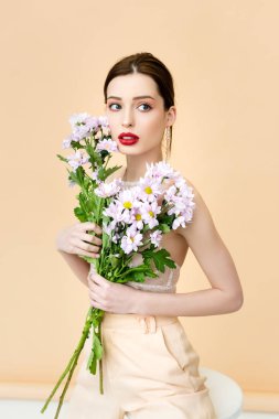 pretty woman looking away and holding blooming chrysanthemum flowers on beige 