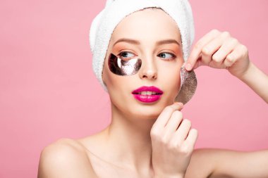 naked young woman in towel applying eye patches isolated on pink 