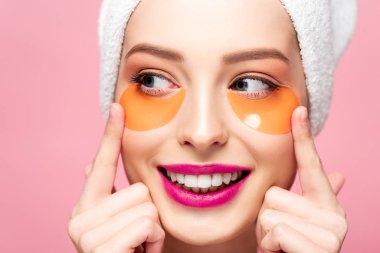 smiling girl touching face with eye patches and looking away isolated on pink 