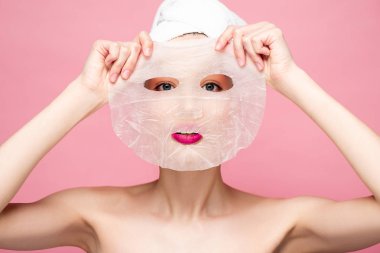 young naked woman covering face while applying moisturizing face mask isolated on pink 