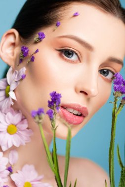 attractive woman looking at camera near blooming flowers isolated on blue 