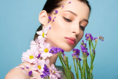 attractive and naked woman looking at blooming flowers isolated on blue 