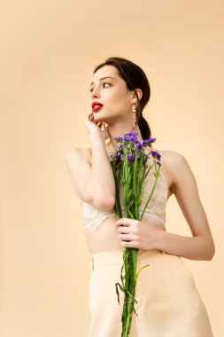 attractive woman holding purple limonium and looking away flowers isolated on beige 