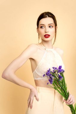 young attractive woman holding purple limonium flowers and standing with hand on hip isolated on beige 
