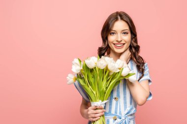 happy girl touching neck while holding bouquet of white tulips and smiling at camera isolated on pink clipart