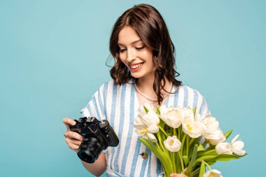 Dijital fotoğraf makinesi ve mavi üzerinde izole edilmiş beyaz laleler tutan neşeli genç bir kadın.