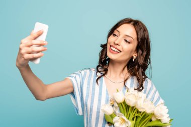 Mutlu genç bir kadın akıllı telefondan selfie çekerken, laleler mavi renkte izole edilmiş.