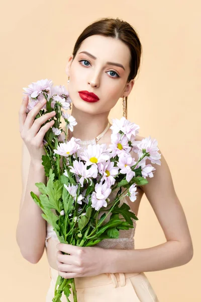 attractive young woman holding blooming flowers and looking at camera isolated on beige 