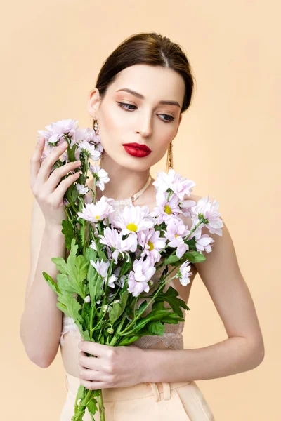 pretty young woman looking away near blooming chrysanthemum flowers isolated on beige 