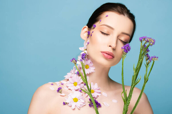 naked young woman with closed eyes near blooming flowers isolated on blue 