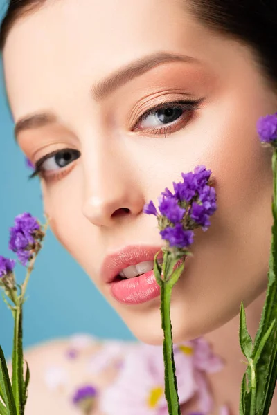 close up of pretty woman looking at camera near blooming flowers isolated on blue 
