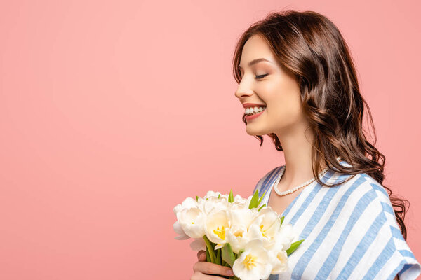 happy girl smiling with closed eyes while holding bouquet of white tulips isolated on pink