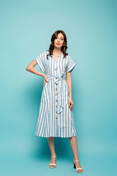 full length view of attractive young woman looking at camera while holding hand on hip on blue background