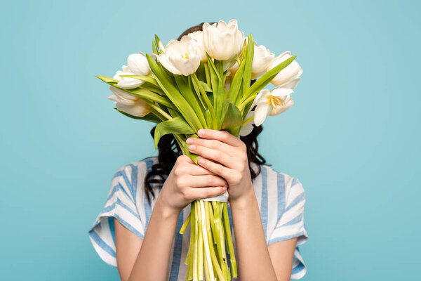 young woman obscuring face with bouquet of white tulips isolated on blue