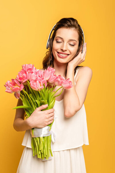 happy young woman holding bouquet of pink tulips while listening music in wireless headphones with closed eyes isolated on yellow