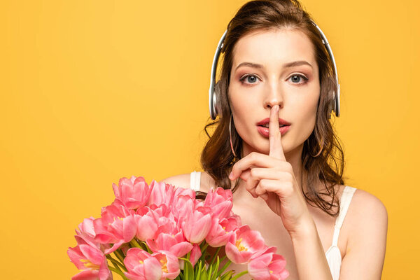attractive young woman in wireless headphones showing hush gesture while holding bouquet of tulips isolated on yellow