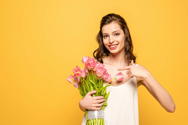smiling young woman pointing with finger at bouquet of pink tulips while looking at camera isolated on yellow