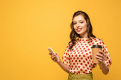 attractive young woman holding coffee to go while using smartphone and looking at camera isolated on yellow