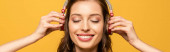 Panoramaaufnahme einer glücklichen jungen Frau, die Musik in drahtlosen Kopfhörern hört, mit geschlossenen Augen, isoliert auf gelb