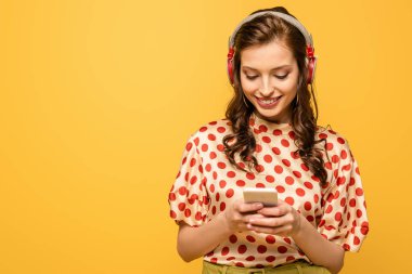 Kablosuz kulaklıklı neşeli genç kadın sarıda izole edilmiş akıllı telefondan sohbet ediyor.