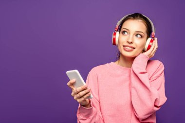 Gülümseyen, hayalperest kız kablosuz kulaklıkla müzik dinlerken elinde akıllı telefon tutarken ve mor renkte izole bir şekilde yüzünü çevirerek uzaklaşıyor.