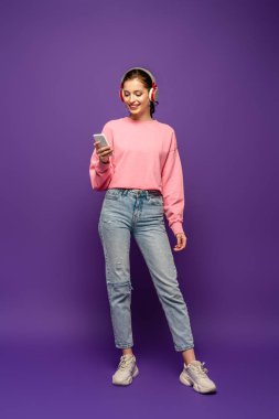 Mor arka planda akıllı telefondan sohbet ederken gülümseyen, kablosuz kulaklıklı güzel kızın tam boy görüntüsü