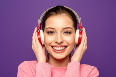 Mor renkte izole edilmiş kablosuz kulaklıklara dokunurken kameraya gülümseyen mutlu kız.