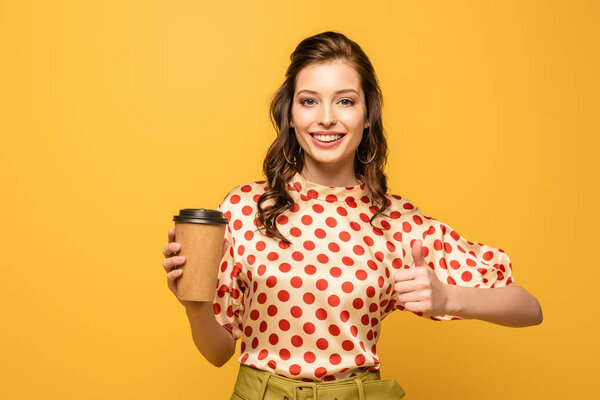happy young woman holding coffee to go and showing thumb up while looking at camera isolated on yellow