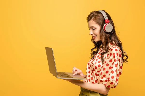 cheerful young woman in wireless headphones using laptop isolated on yellow