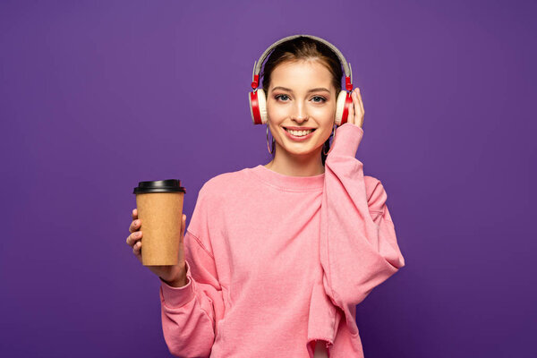 smiling girl holding coffee to go while listening music in wireless headphones isolated on purple