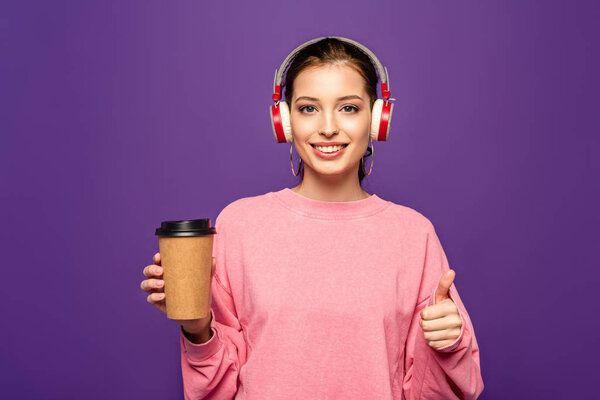 cheerful girl holding coffee to go while listening music in wireless headphones isolated on purple
